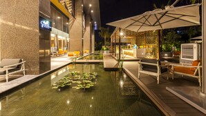 Un área de piscina nocturna con sillones de descanso, una pequeña fuente con nenúfares y un restaurante con mesas al aire libre.