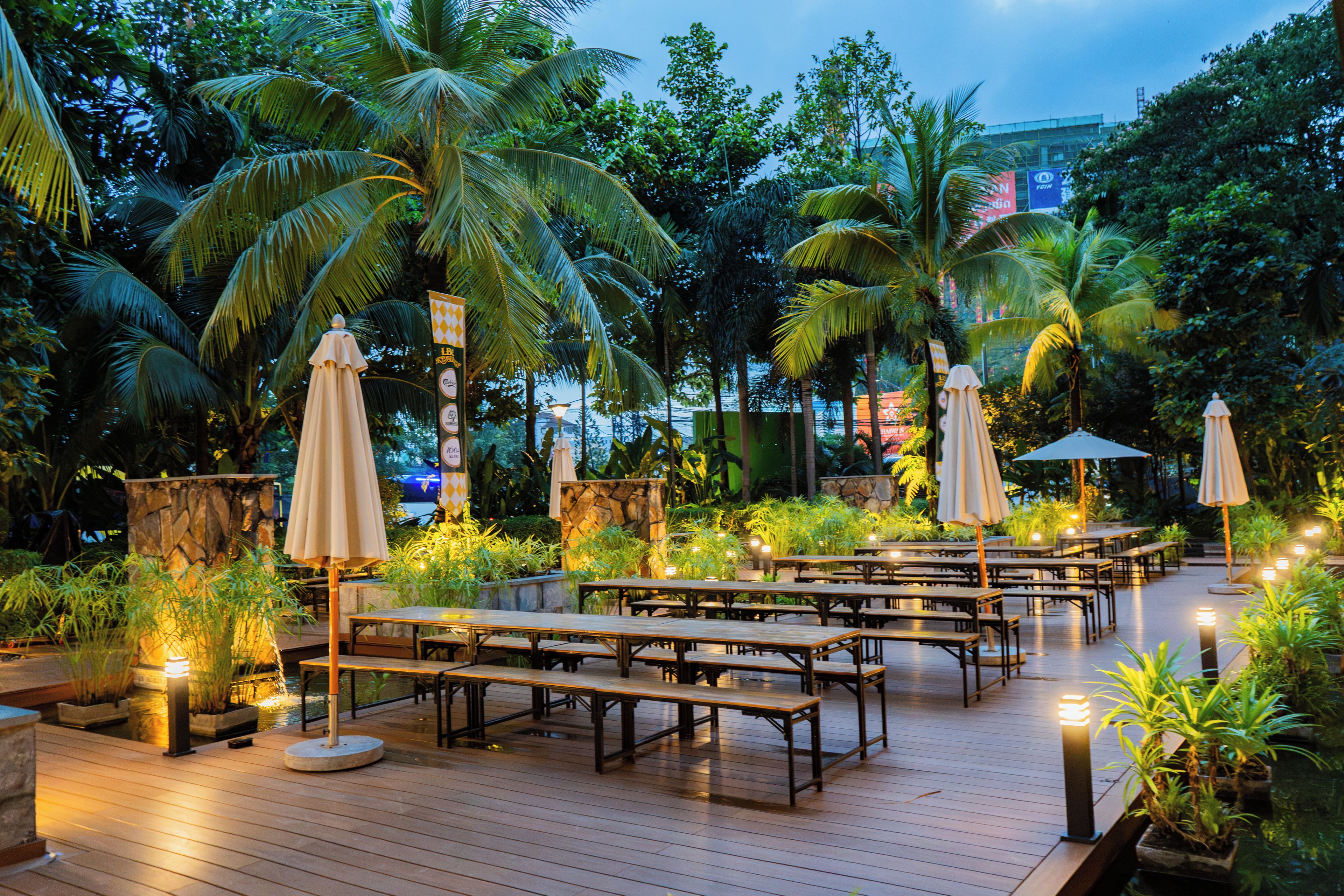 An outdoor dining area with wooden tables and benches, surrounded by lush greenery and palm trees.