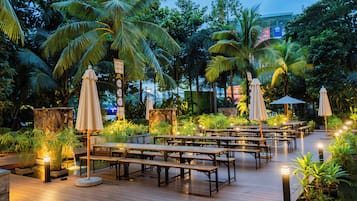 An outdoor dining area with wooden tables and benches, surrounded by lush greenery and palm trees.
