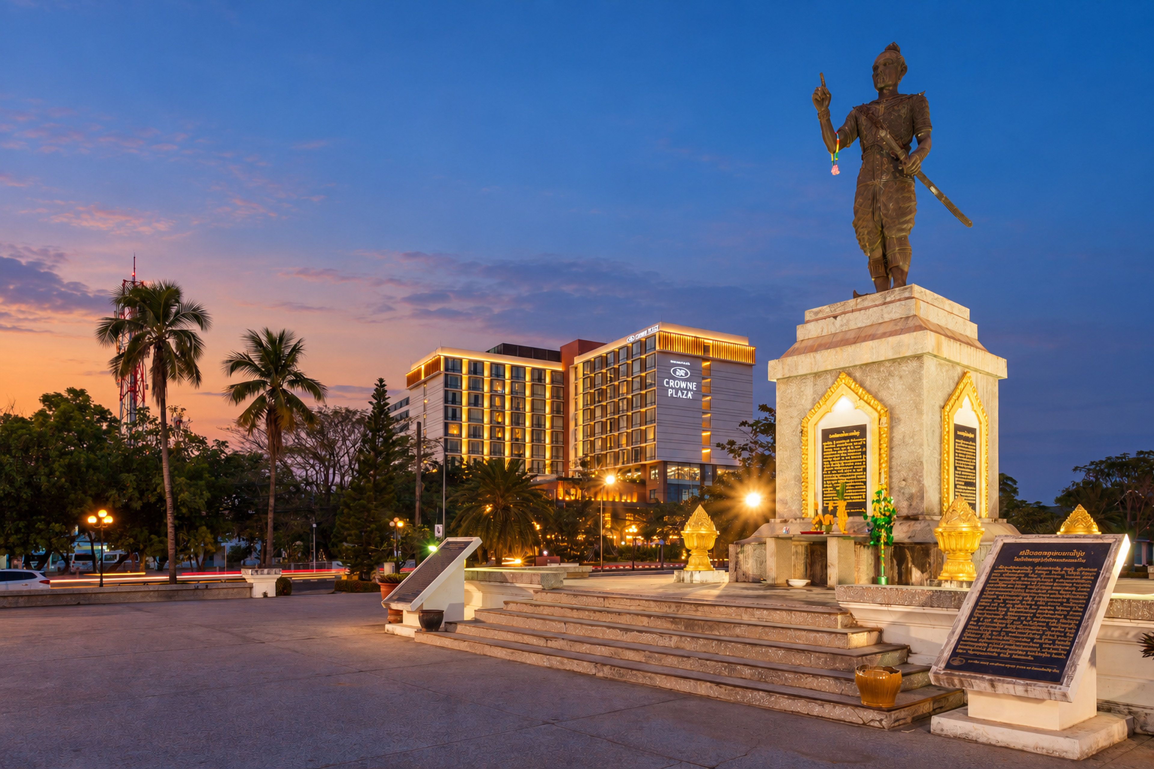 A statue in a public square with steps leading up to it, palm trees, and modern buildings in the background.