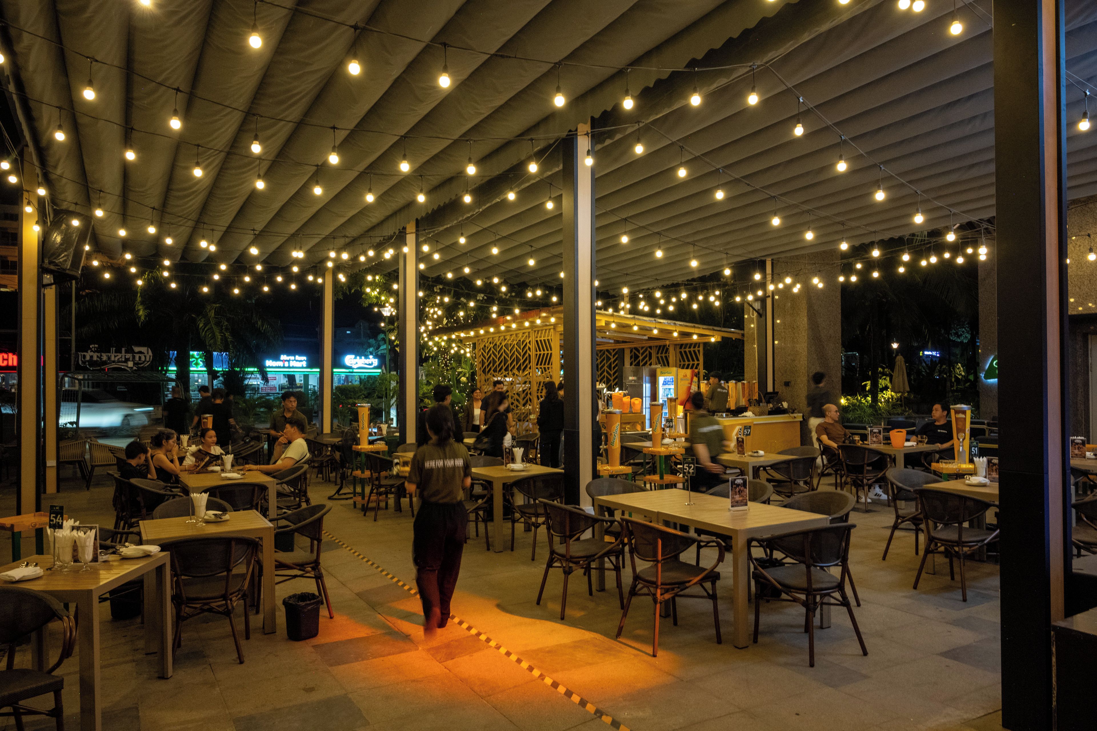An outdoor dining area with tables and chairs, string lights overhead, and a central walkway.