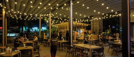 An outdoor dining area with tables and chairs, string lights overhead, and a central walkway.