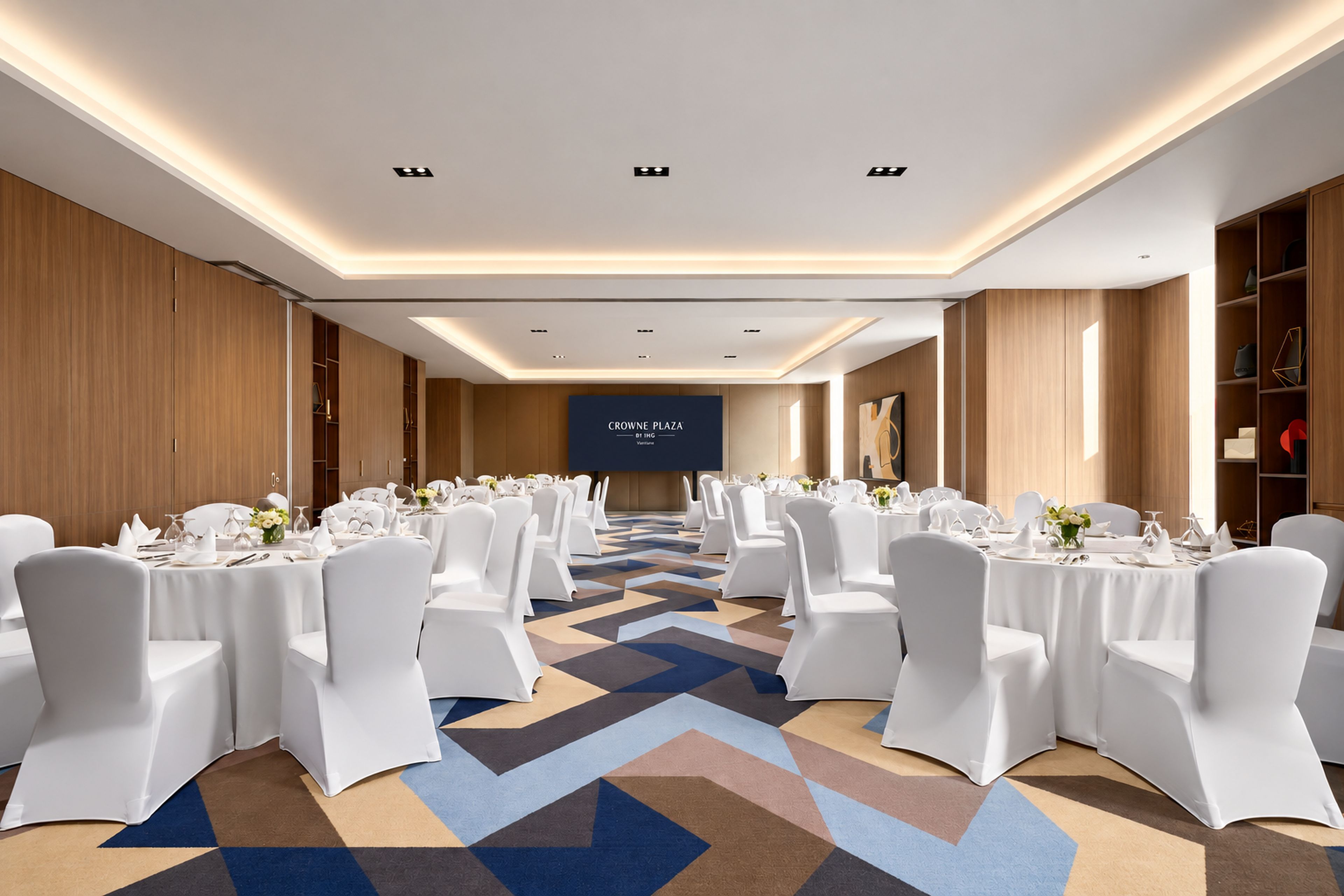 A conference room with white chairs, tables set for dining, and a large screen displaying 'CROWNE PLAZA DT IHG'.