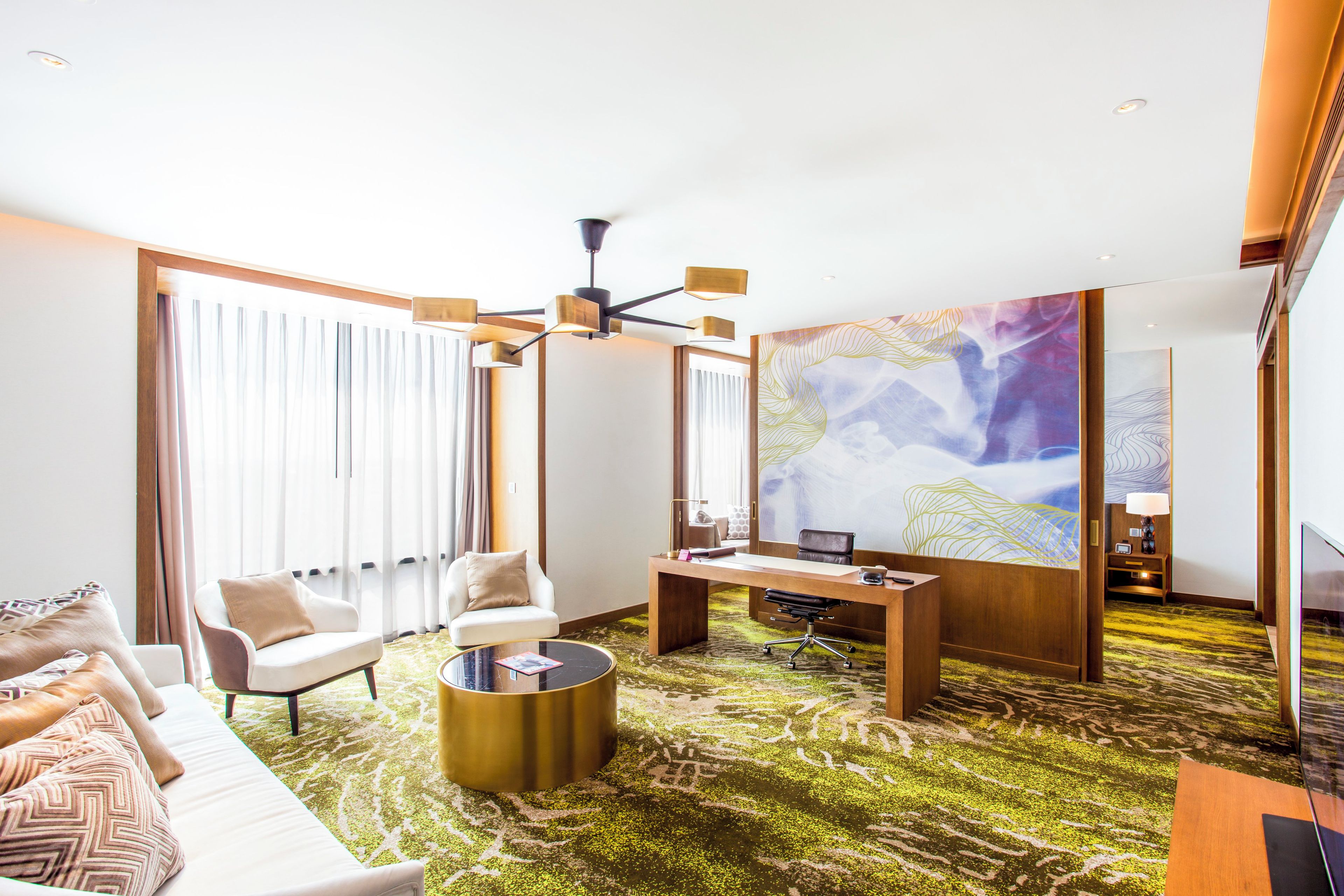 A modern hotel room with a sofa, armchair, desk, and a large abstract painting.
