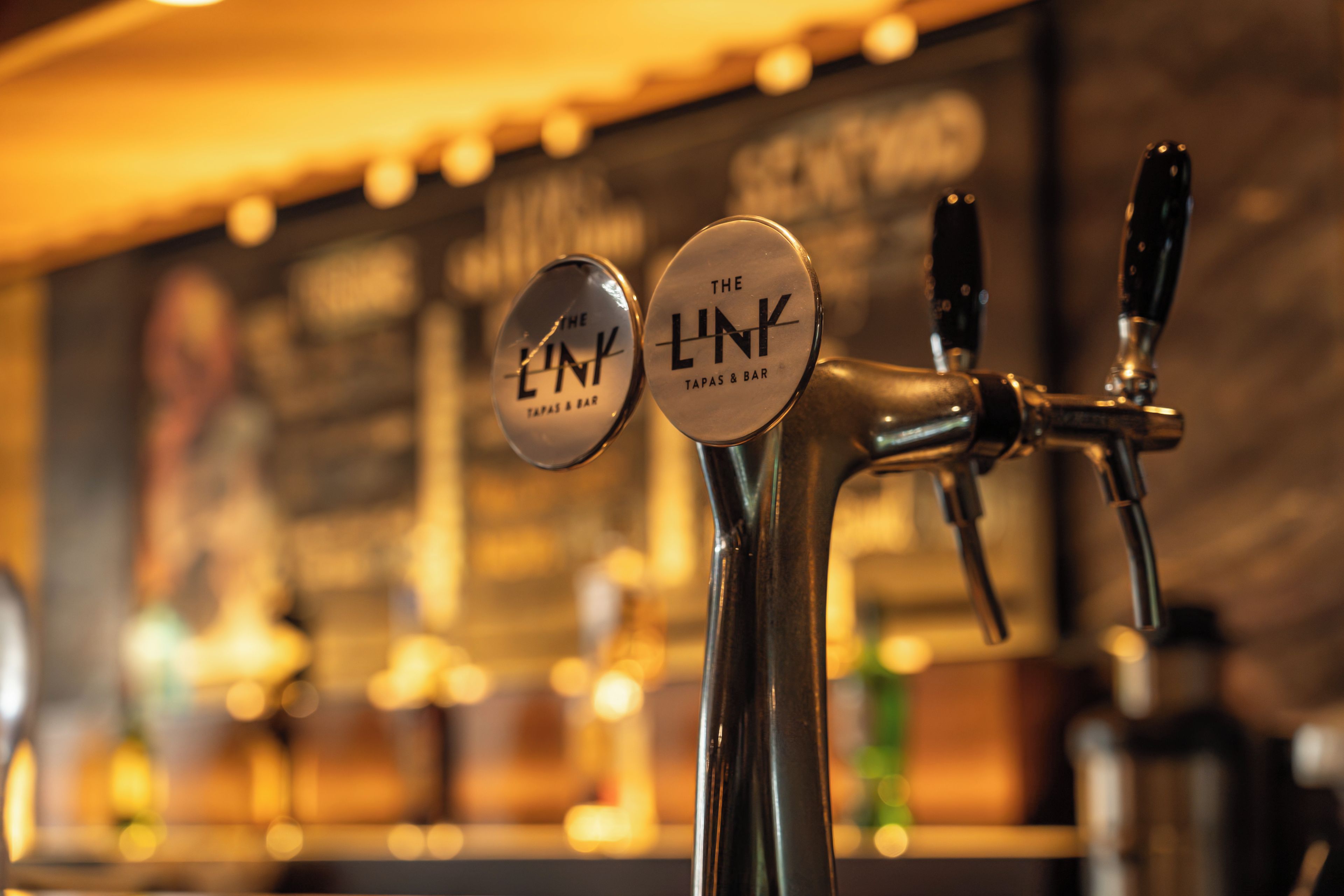 Un bar con due rubinetti, uno dei quali reca l'indicazione 'The Link Tapas & Bar', e uno sfondo sfocato con luci.