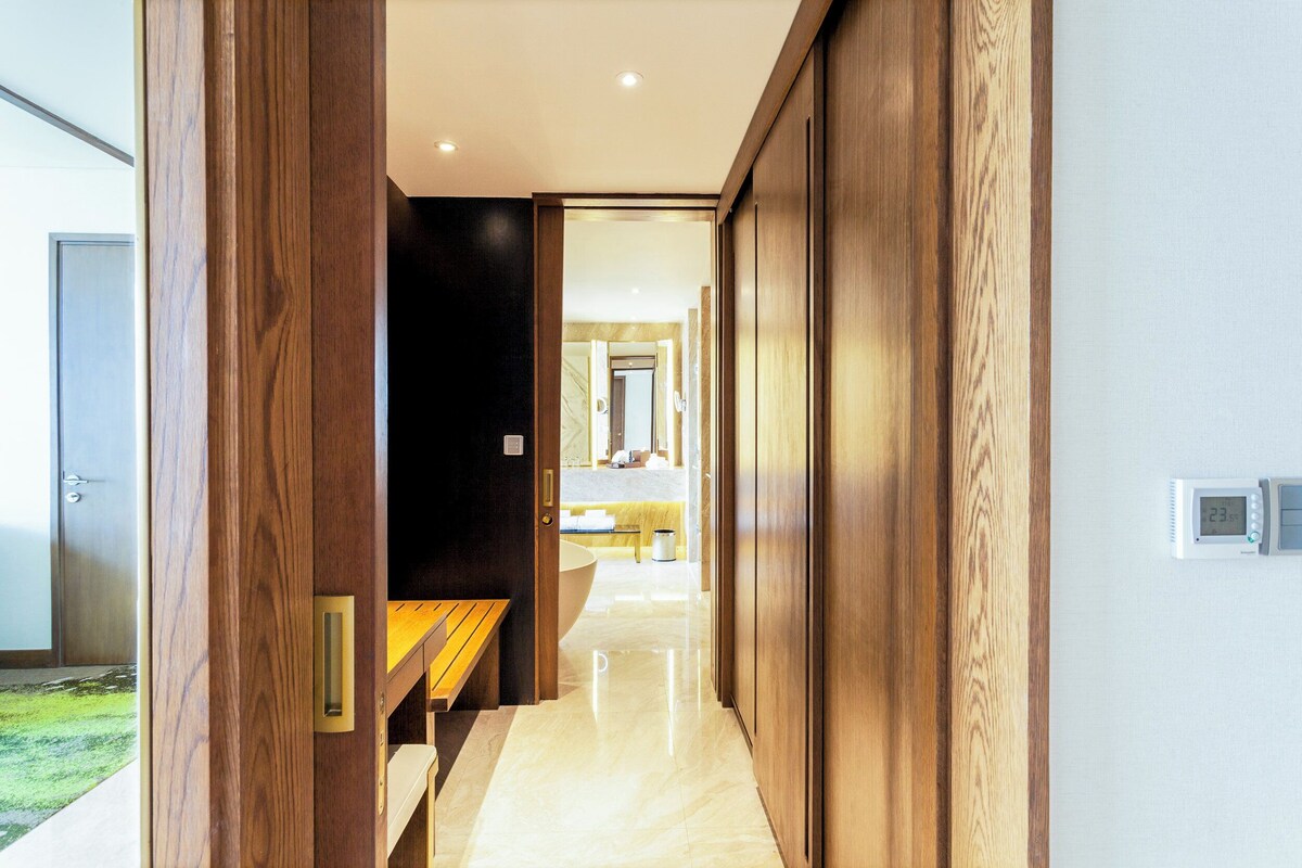 A modern hotel hallway with wooden paneling, a bench, and a view into a well-lit room with a bed and bathroom.