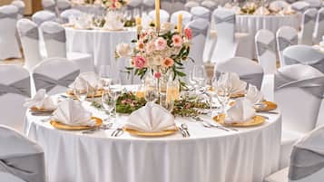 Un salón de banquetes con mesas redondas dispuestas para un evento formal, con manteles blancos, cubertería dorada y centros de mesa florales.