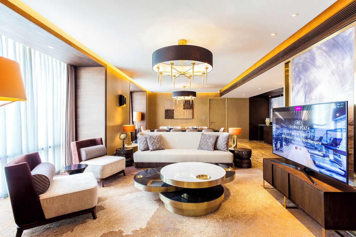 A modern hotel room with a large flat-screen TV, a sofa set, a coffee table, and a chandelier.