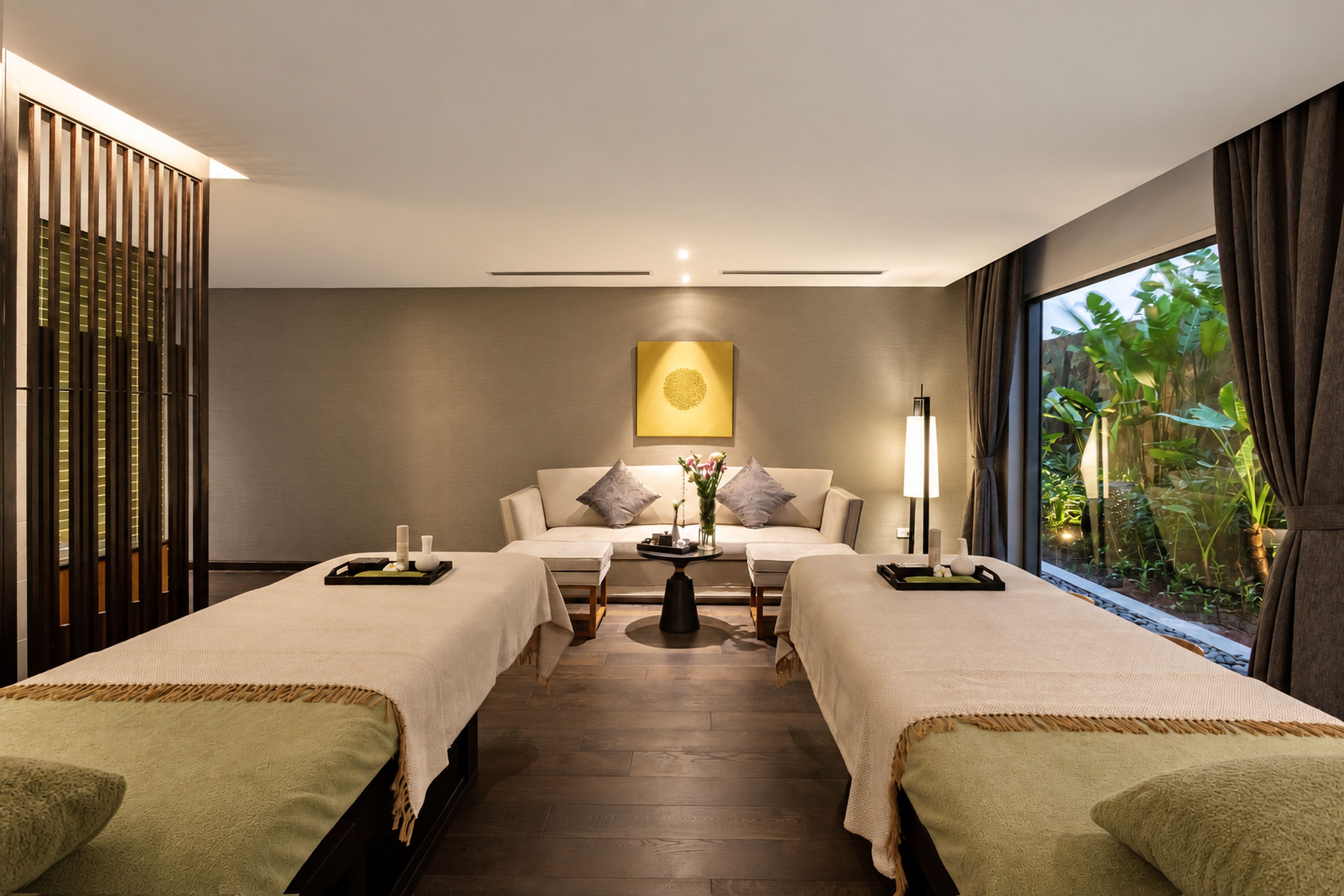 A modern spa room with two massage tables, a central seating area, and a large window with a view of greenery.