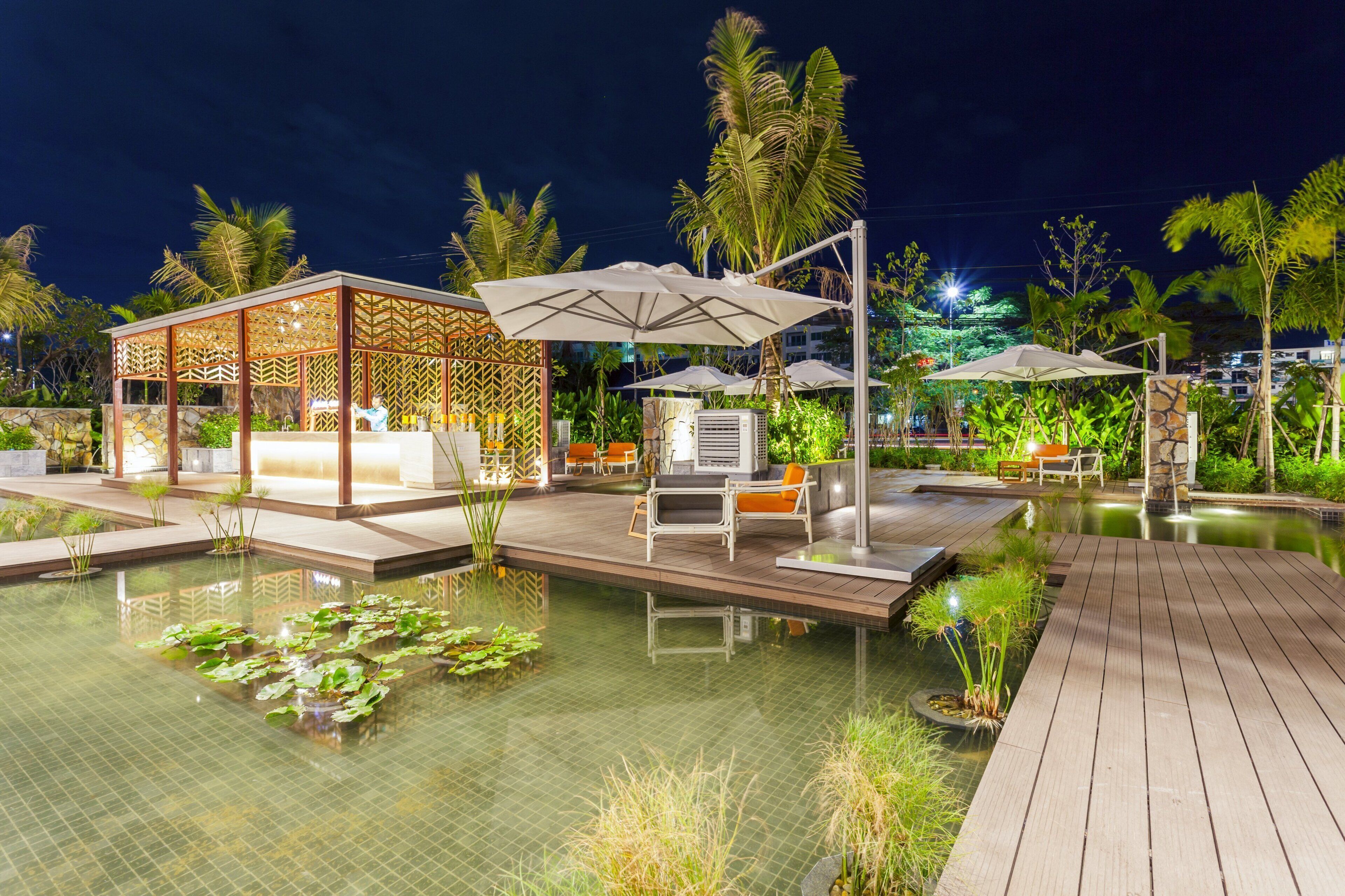 A nighttime resort pool area with a wooden deck, lounge chairs, umbrellas, and a covered seating area.