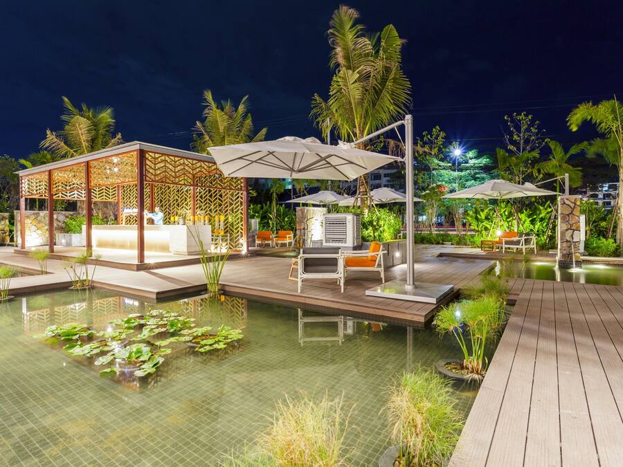 Un área de piscina en un complejo turístico de noche, con muelle de madera, sillas de descanso, sombrillas y un espacio de estar cubierto.