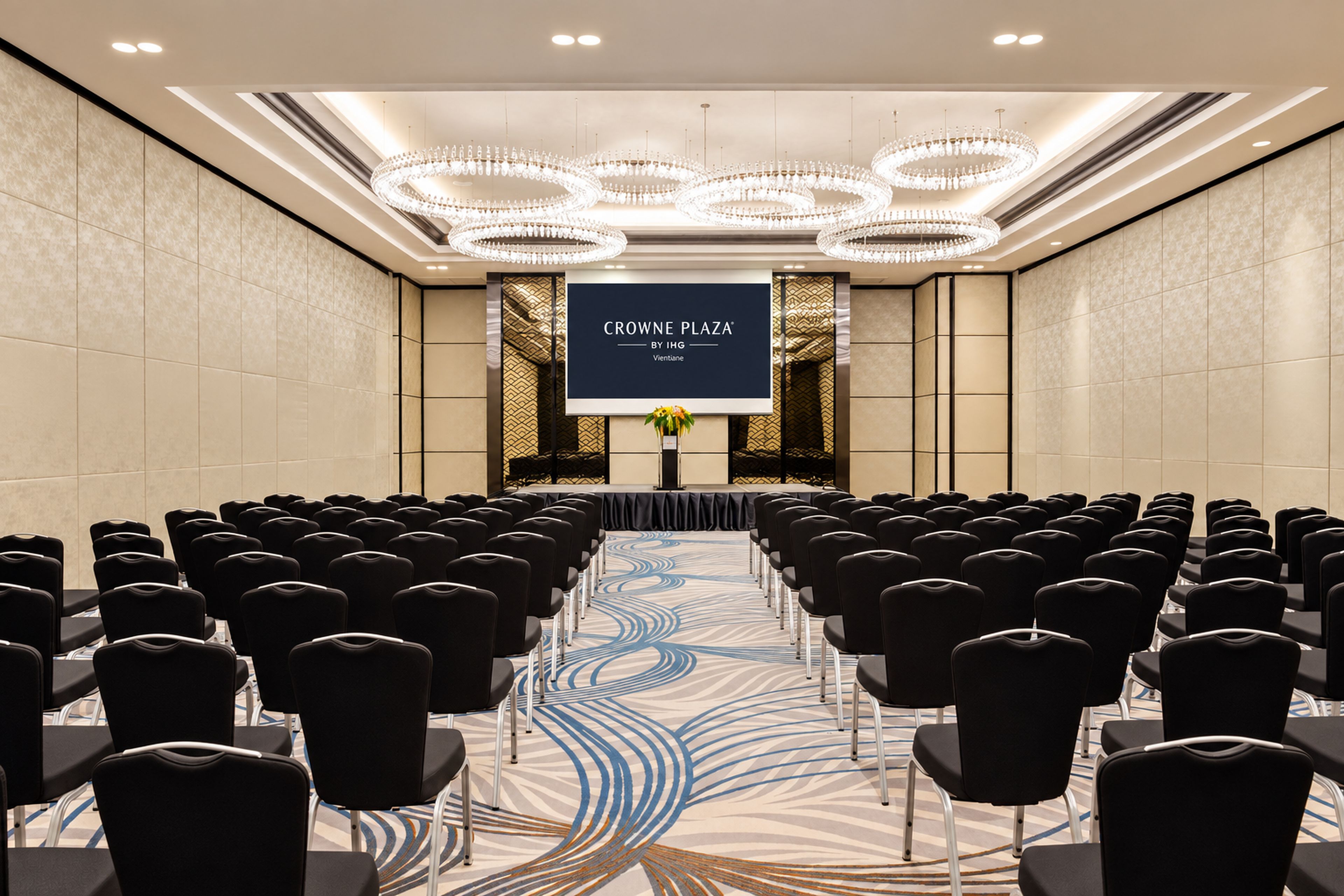 A conference room with rows of chairs, a stage with a screen displaying 'CROWN PLAZA BY IHG', and a patterned carpet.
