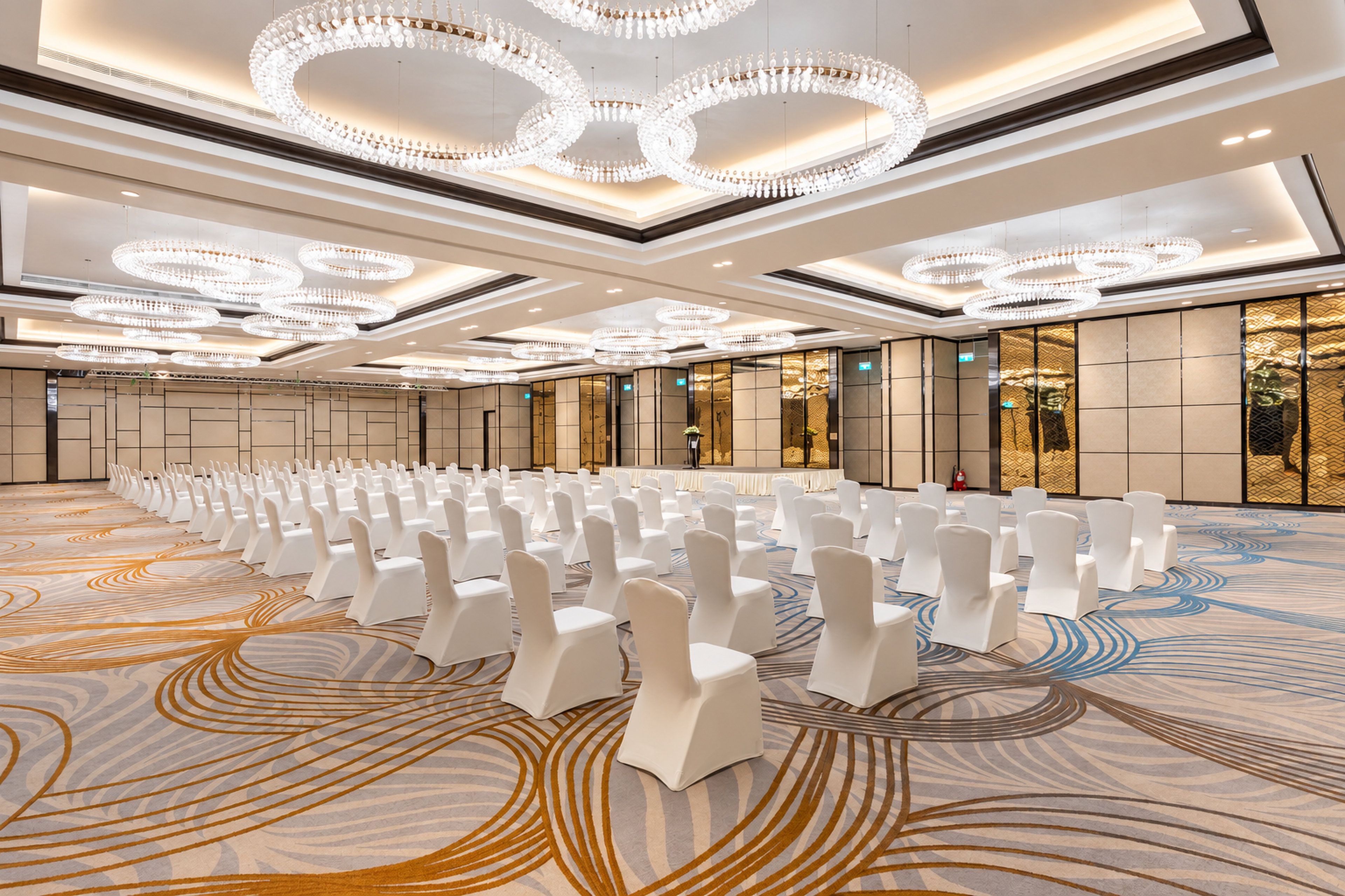 A large hall with rows of white chairs, ornate chandeliers, and patterned carpet flooring.