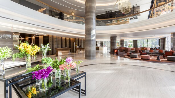 Un vestíbulo amplio con una mesa de vidrio central que exhibe arreglos florales, una escalera moderna y un área de estar con sofás y una mesa de centro.