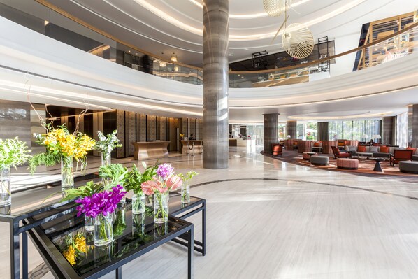 Un vestíbulo amplio con una mesa de vidrio central que exhibe arreglos florales, una escalera moderna y un área de estar con sofás y una mesa de centro.