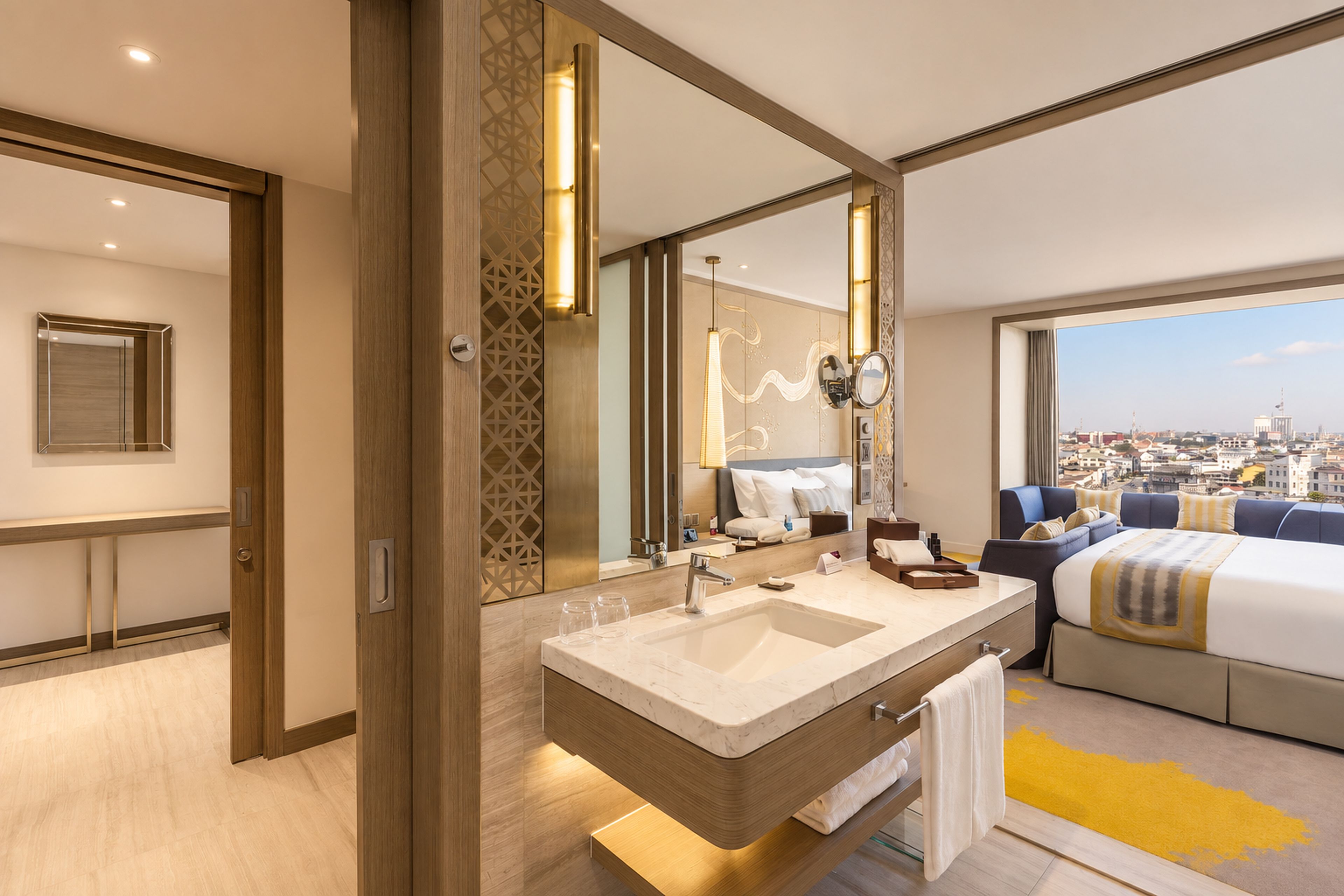 A modern hotel room with a large bed, a bathroom with a double sink, and a city view.