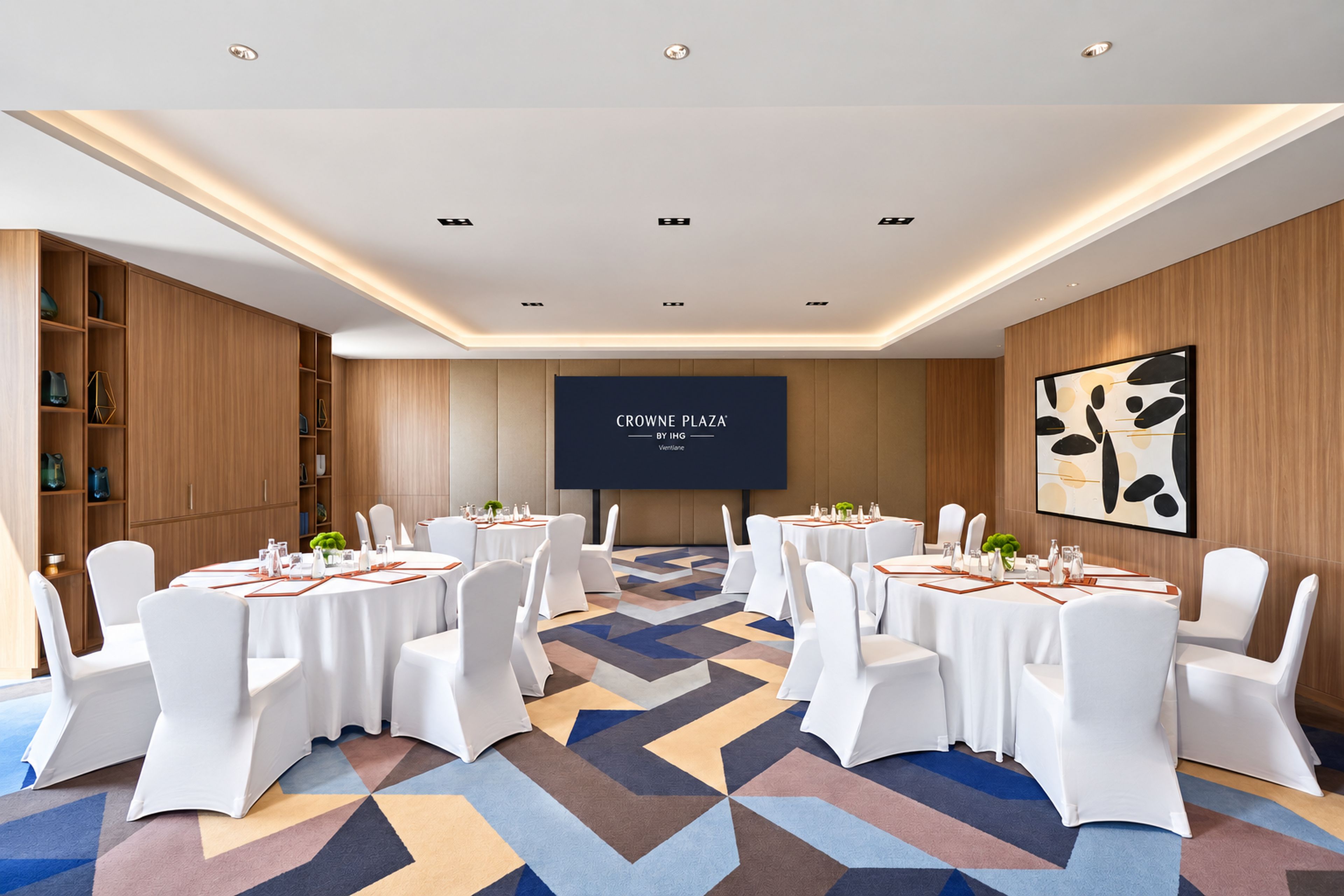 Une salle de conférence équipée de tables rondes, de chaises blanches et d’un grand écran affichant le logo « CROWN PLAZA BY IHG ».