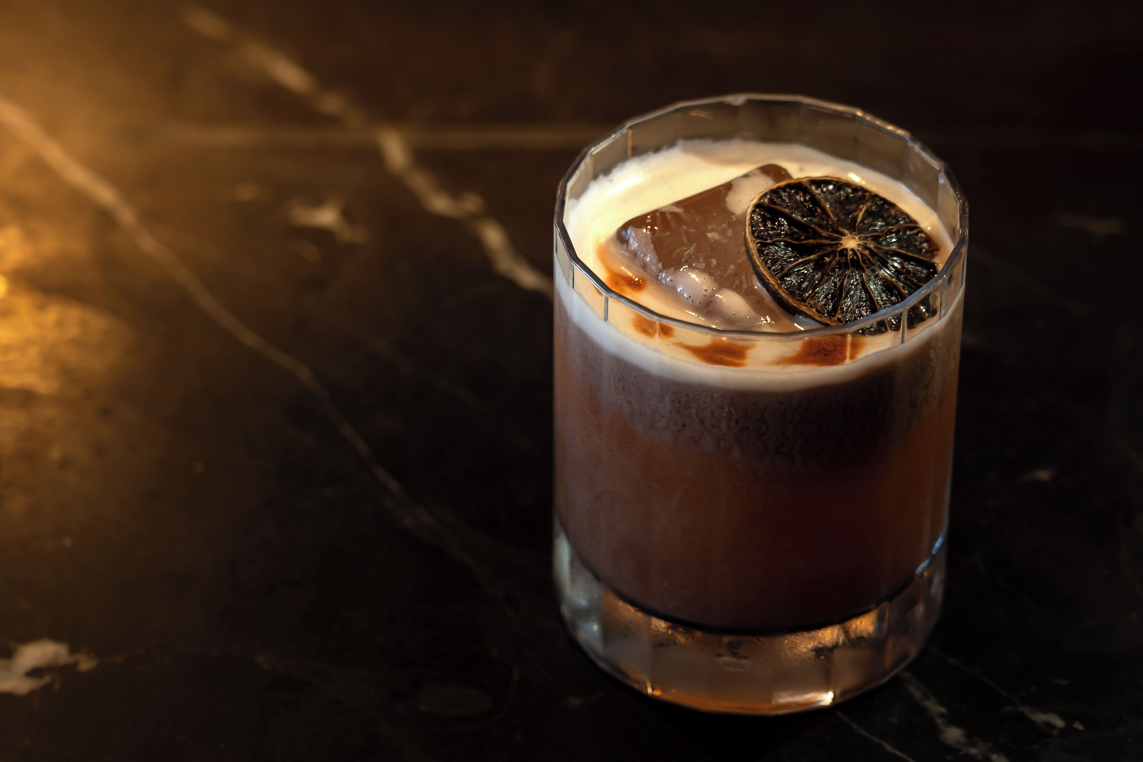 A glass with a dark liquid, ice, and a slice of dried fruit on a dark surface.