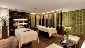 A modern spa room with two beds, a massage table, and a seating area.
