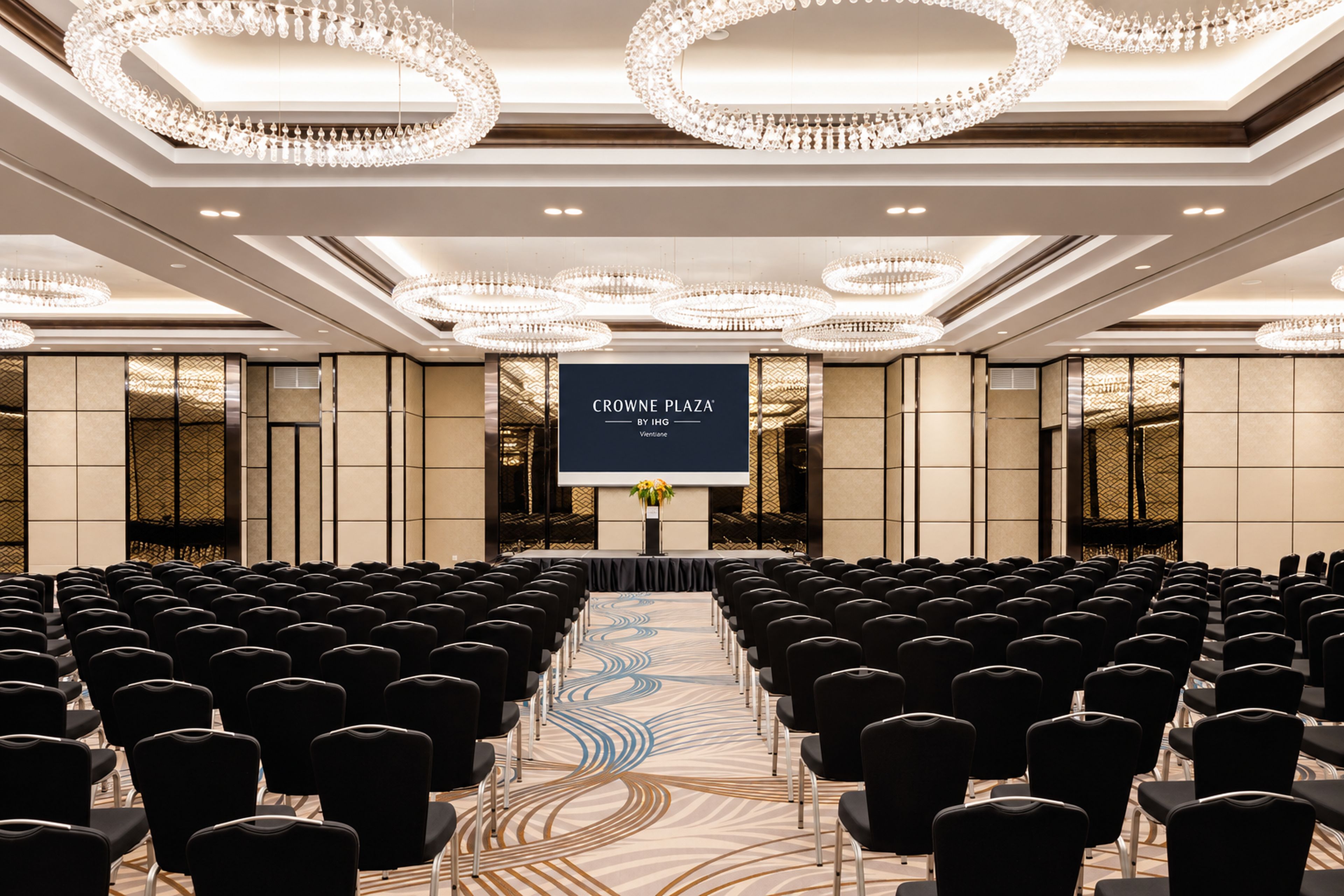 A conference room with rows of chairs facing a stage, a large screen, and ornate chandeliers.