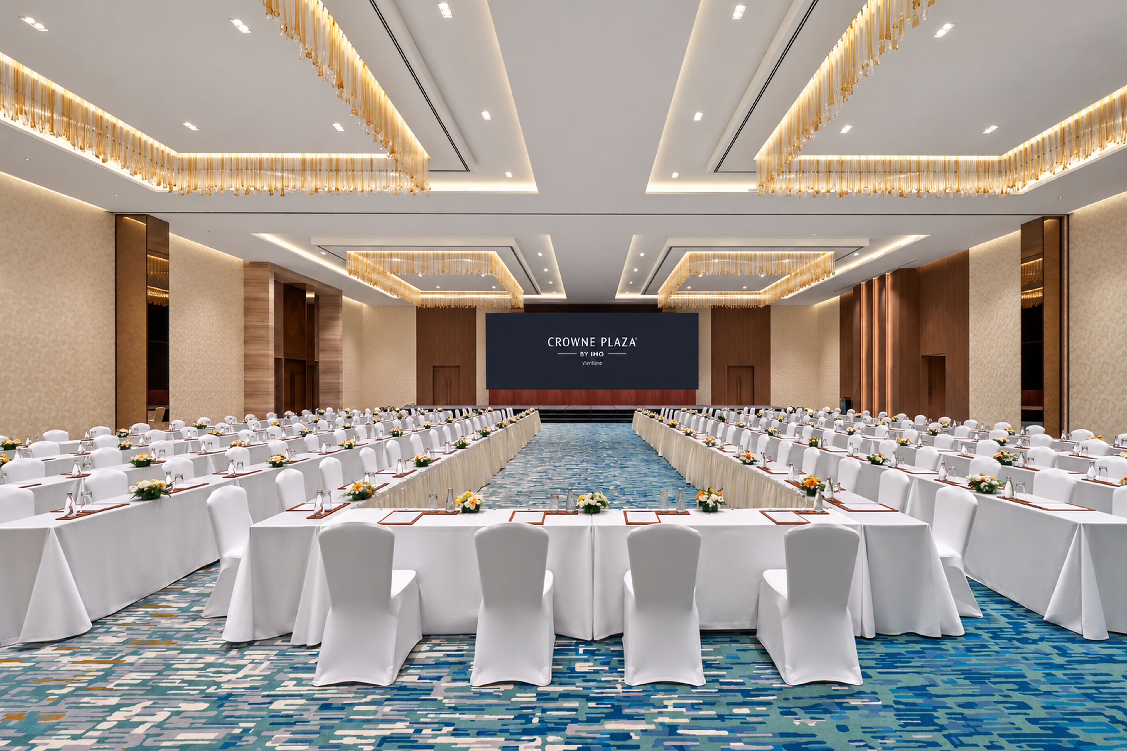 A conference room with rows of tables, white chairs, and a large screen at the front.