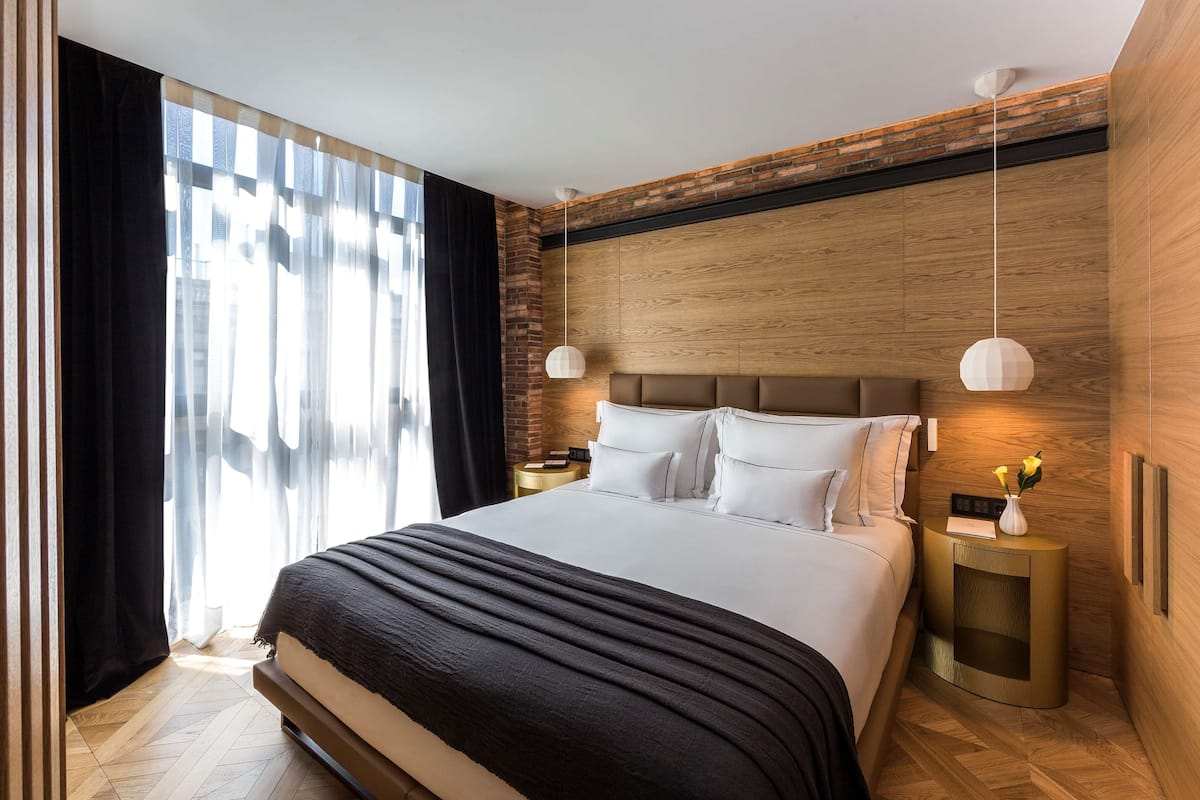 Una habitación de hotel moderna con una cama grande, paneles de madera, una pared de acento de ladrillo y vistas a la ciudad.