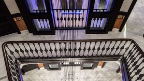 Una imponente escalera con un candelabro central, barandillas ornamentadas y vistas a un edificio con ventanas arqueadas.