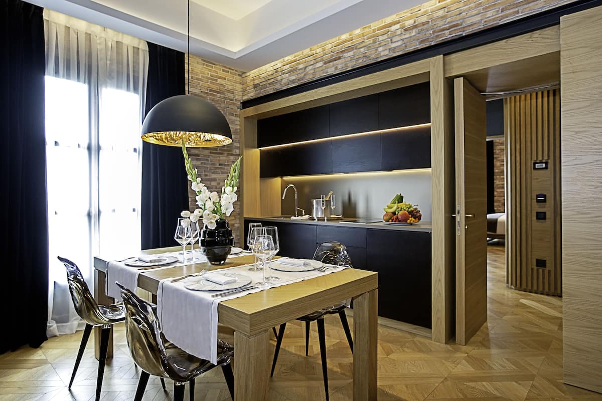 Un comedor moderno con una mesa de madera dispuesta para dos personas, una lámpara colgante negra y una zona de cocina con un frutero a la vista.