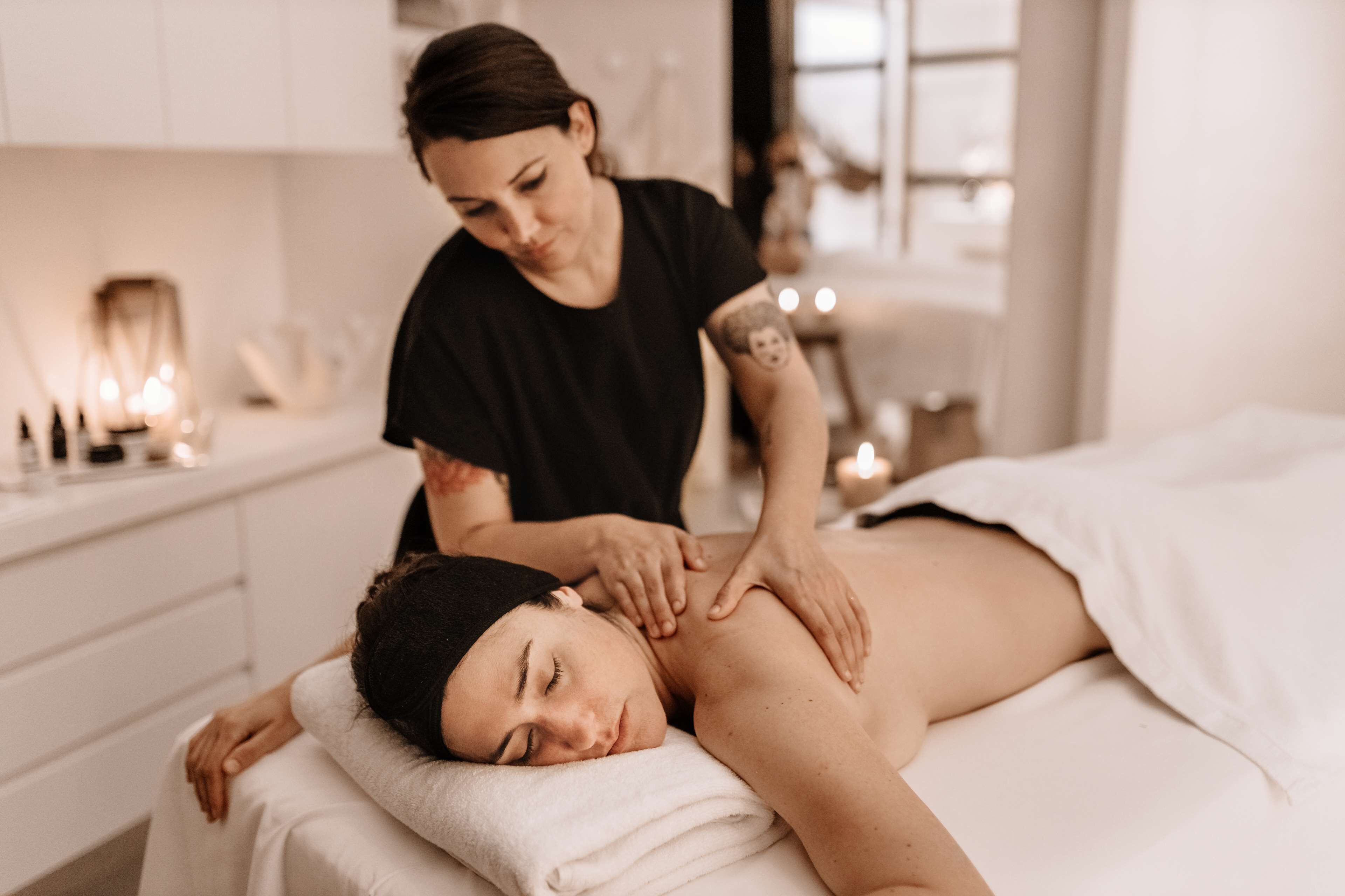 A massage therapy session in a spa setting with a client lying on a treatment table, a therapist attending to the client, and a serene, well-lit room with a window and candles.