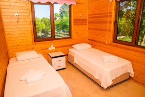 A wooden cabin with two beds, a small bedside table, and large windows offering a view of greenery outside. - Ikiz Villas (Kemer)