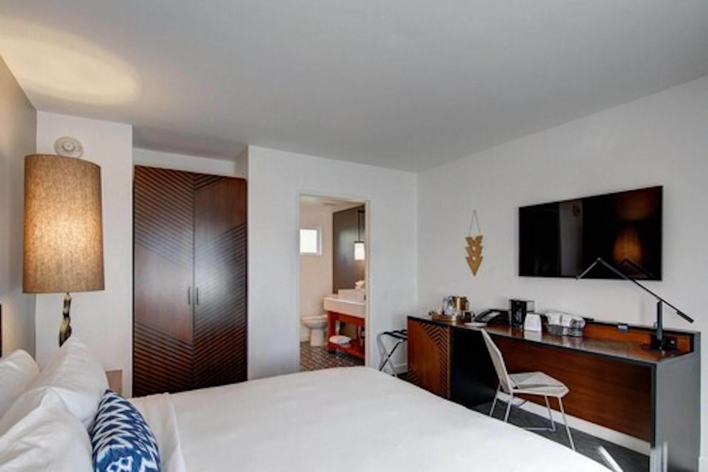 A hotel room with a bed, desk, chair, lamp, wardrobe, and a TV on the wall.