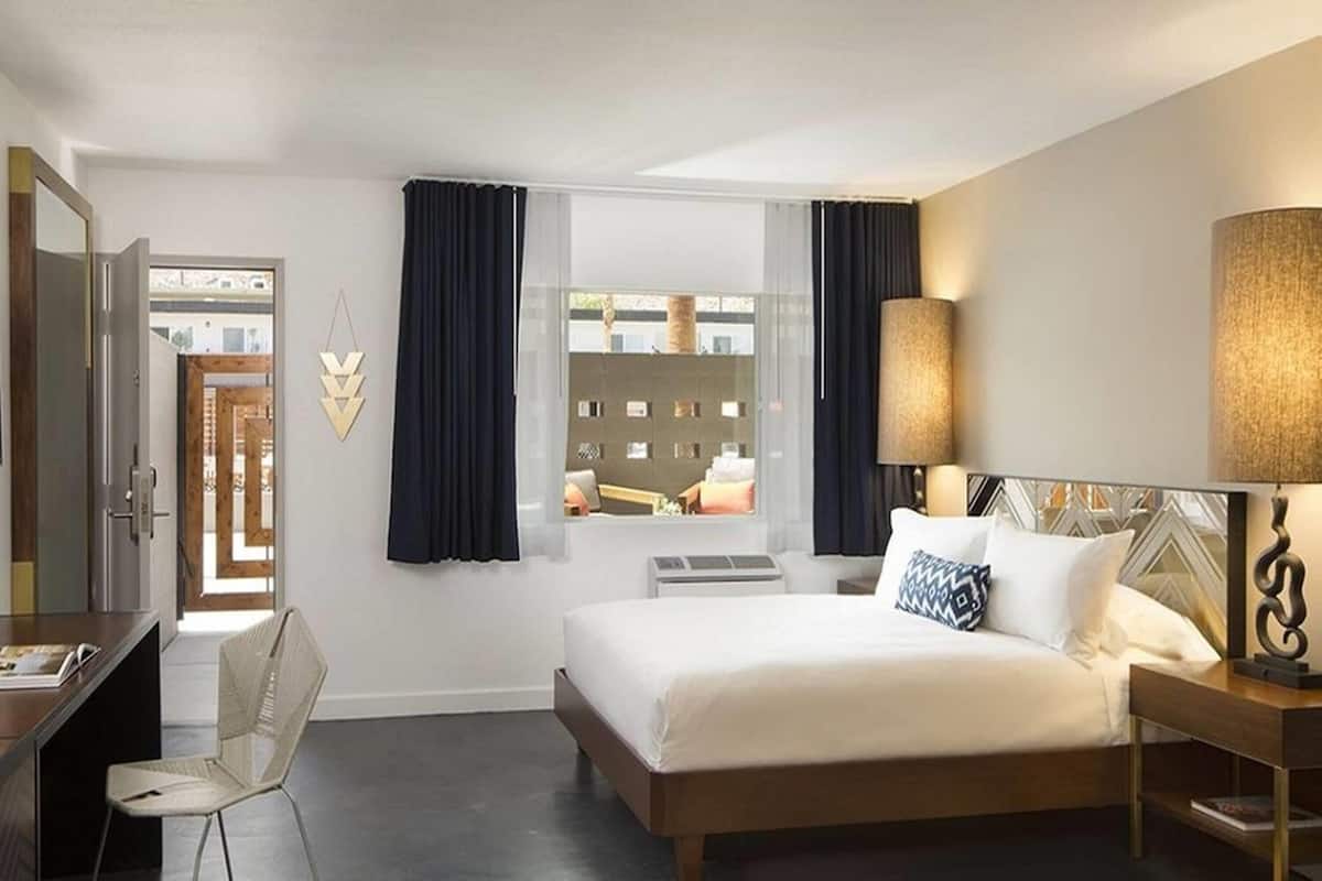 Habitación de hotel con cama, mesita de noche, silla, ventana con cortinas y vistas al exterior.