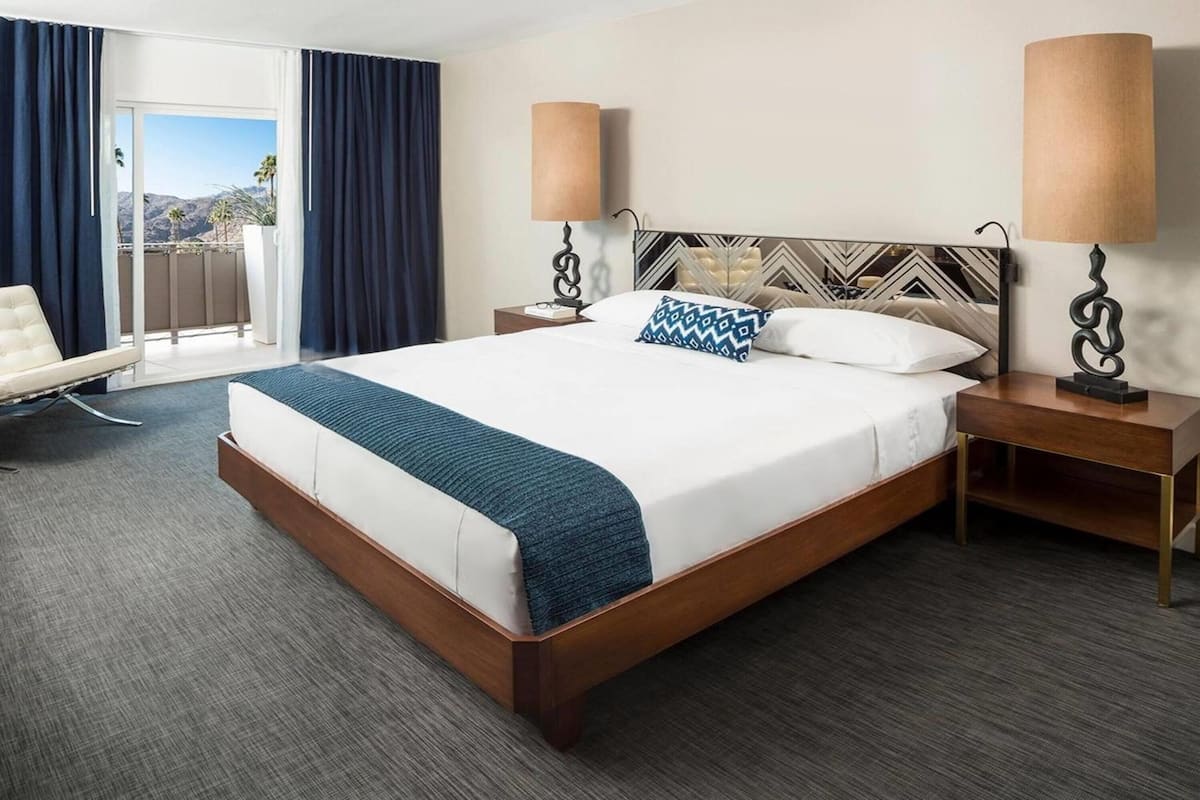 Habitación de hotel con una cama grande, dos mesitas de noche, una silla y vistas al exterior.