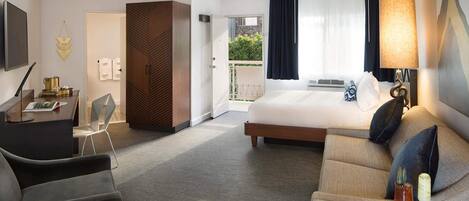 A hotel room with a bed, a sofa, a desk, and a chair. There is a window with curtains and a view of greenery outside.