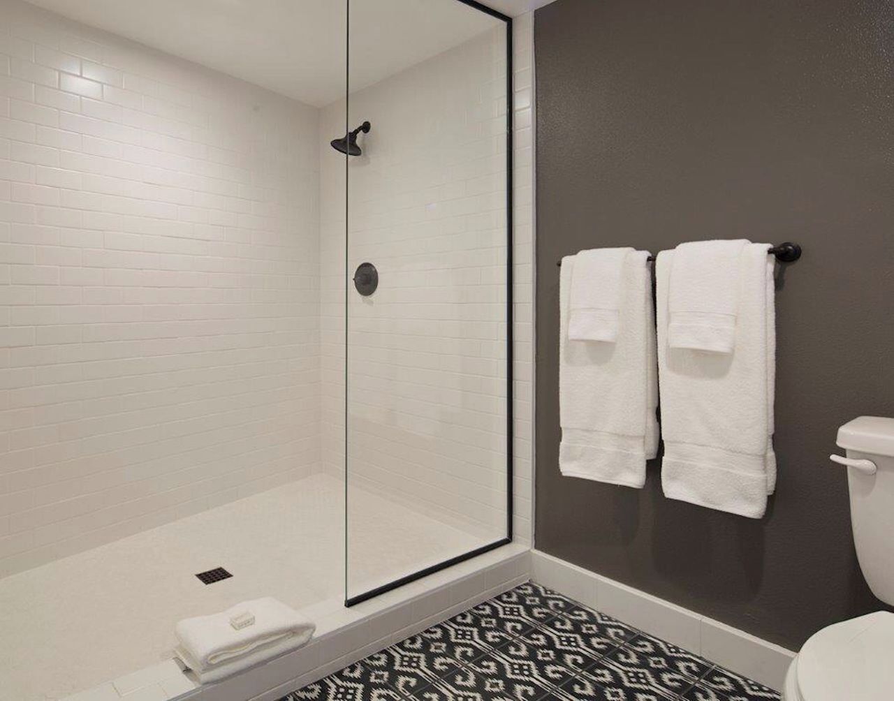 A bathroom with a glass-enclosed shower, white tiled walls, a patterned floor, and two white towels hanging on a rack.