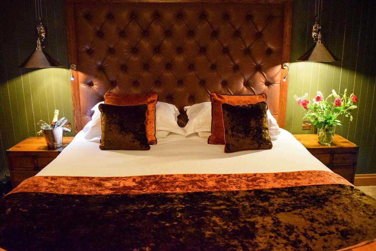 A hotel room with a tufted headboard, a large bed with two pillows, a bedside table with a lamp, and a vase with flowers.