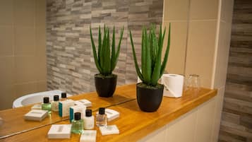 Une salle de bain avec un meuble-lavabo en bois, des plantes d’aloe vera, des articles de toilette et un lavabo blanc.