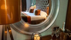 A hotel room with a large round mirror, a bed with multiple pillows, a bedside table with a bottle of champagne, and two empty glasses. - Craigmhor Lodge & Courtyard (Pitlochry)