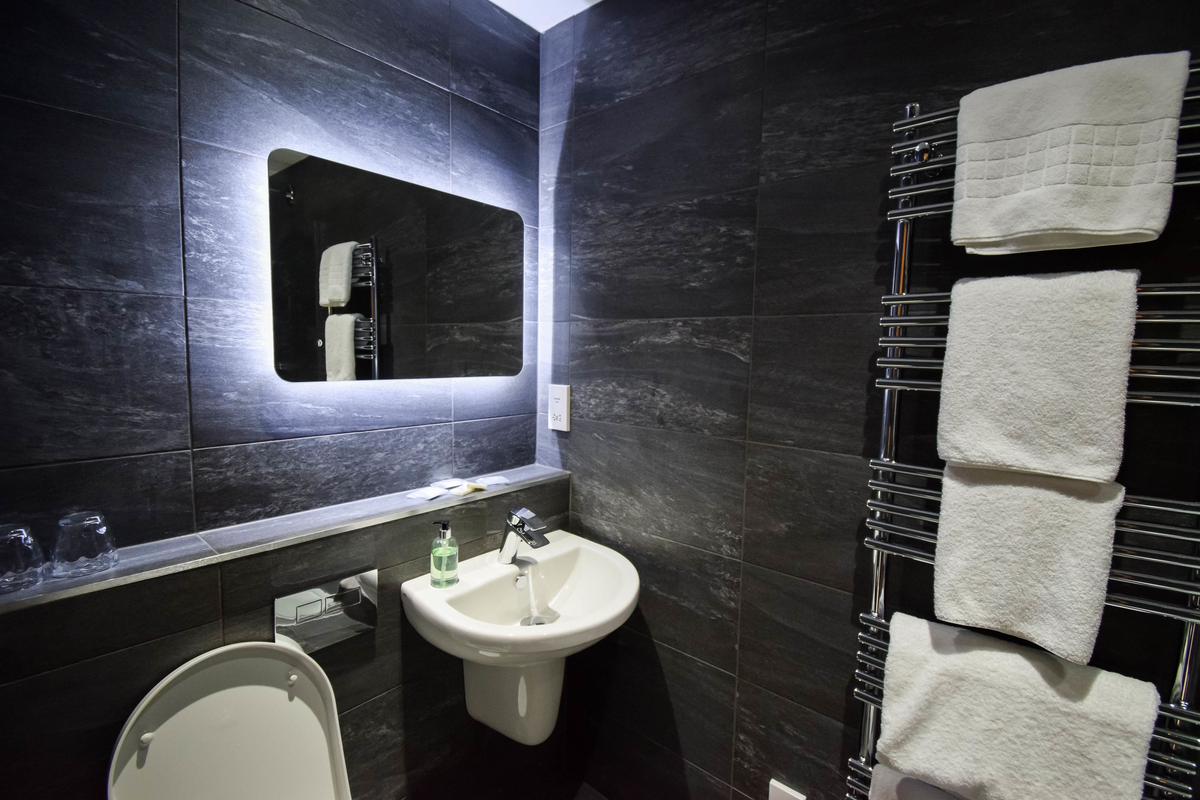 A modern bathroom with a dark tiled wall, a white sink, a mirror, and a towel rack with several towels.