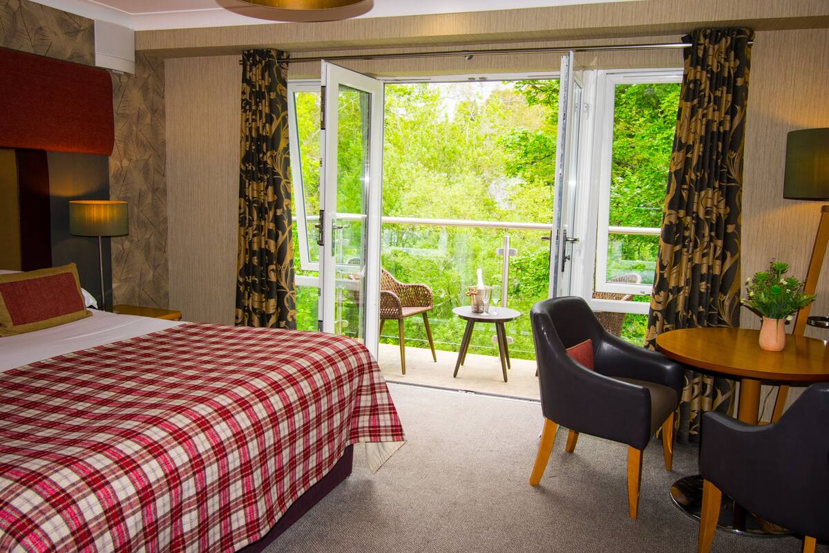 A hotel room with a bed, a desk, a chair, a small table, and a balcony with a view of greenery.