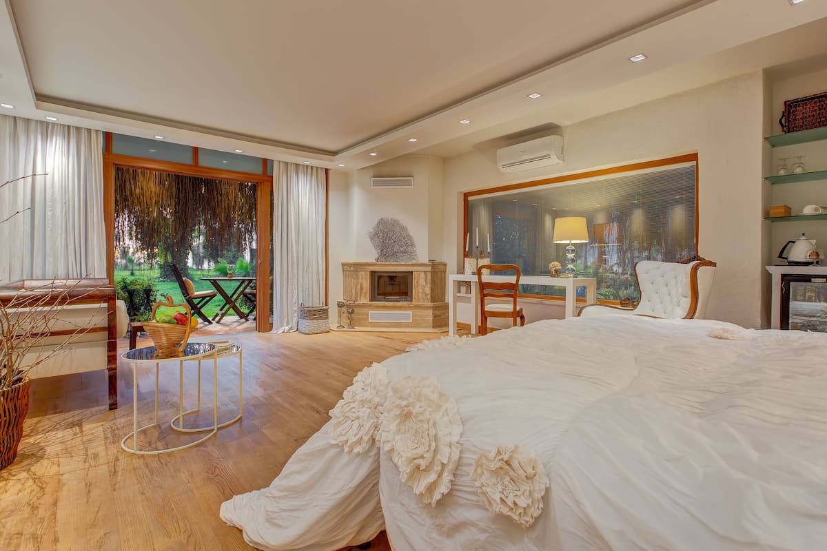 Quarto espaçoso com uma cama grande, piso de madeira, vista para um jardim e um espaço de jantar com mesa e cadeiras.