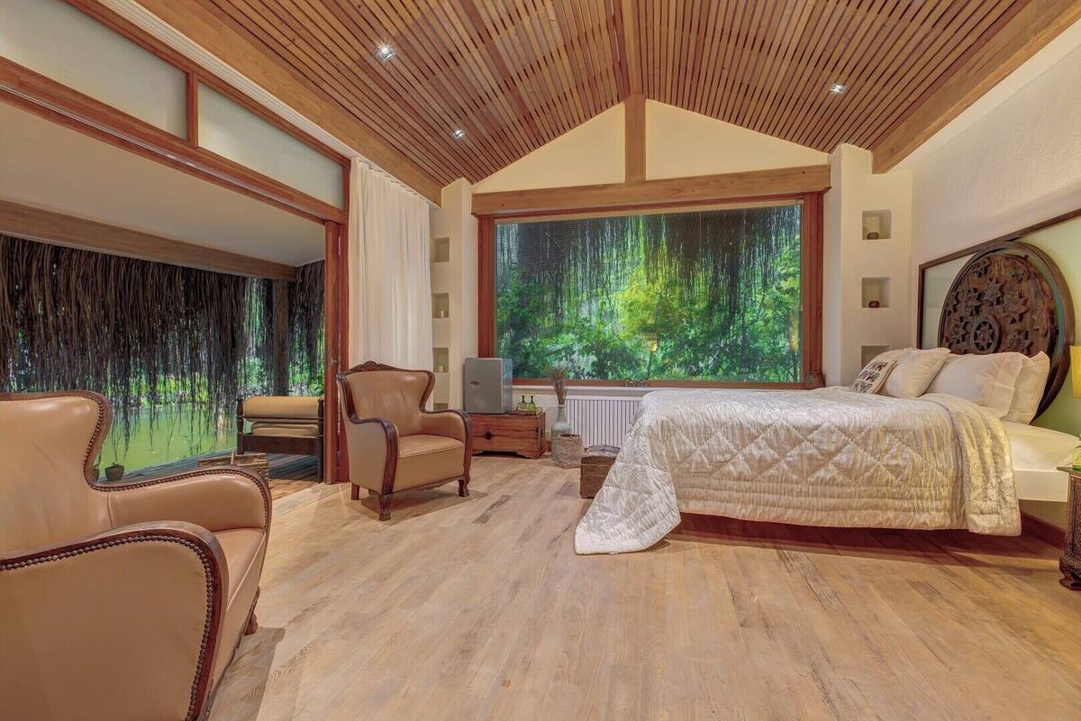 Quarto com uma cama grande, teto de madeira e vista para uma floresta através de uma janela ampla.