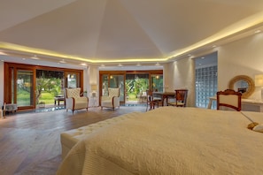Une chambre spacieuse avec un grand lit, deux fauteuils, un bureau et une vue sur un jardin luxuriant, grâce à de grandes fenêtres.