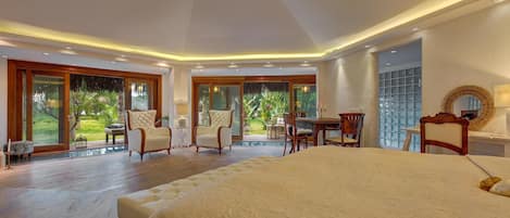A spacious bedroom with a large bed, a desk, and a chair. There is a view of a garden through the windows.