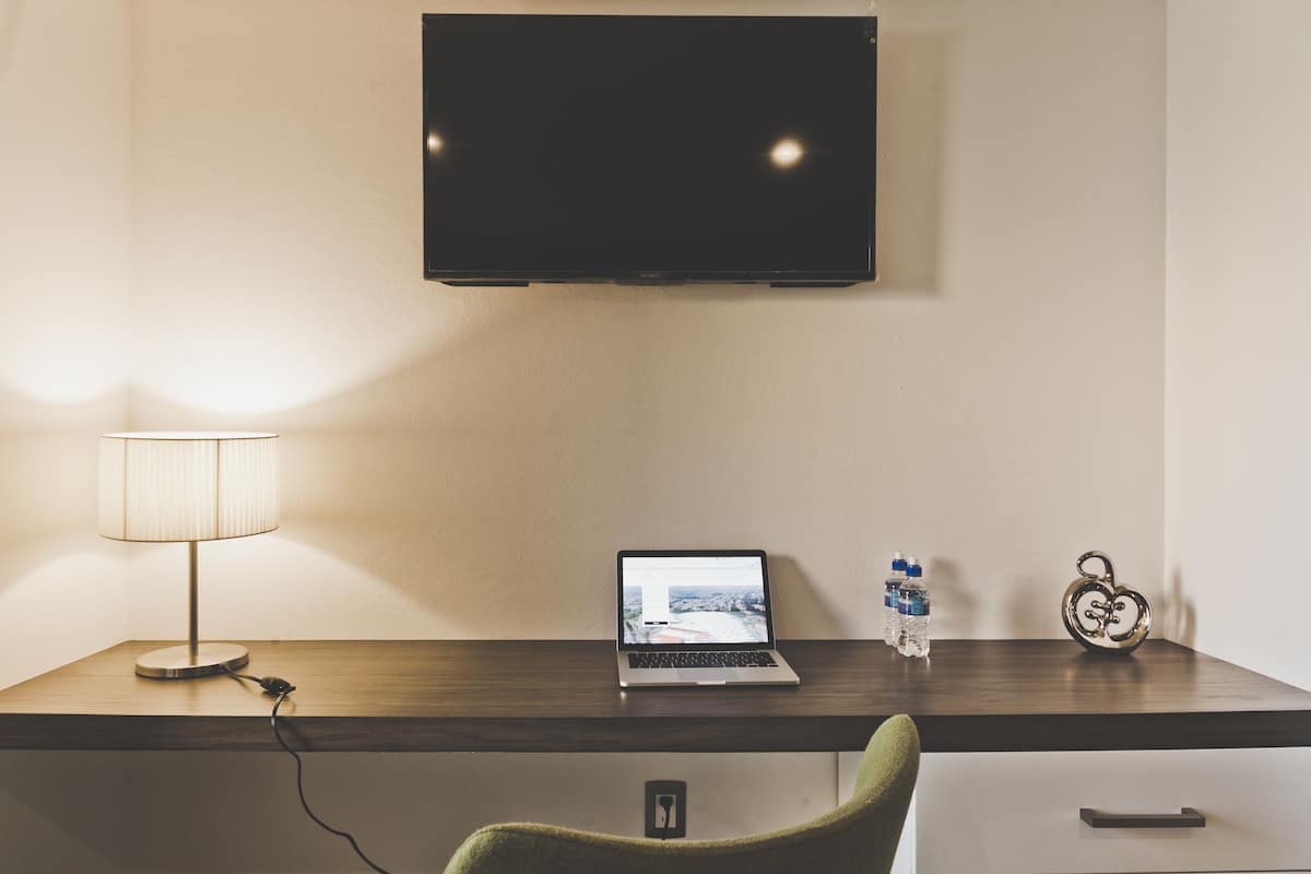 Hotellihuone, jossa on puinen työpöytä, kannettava tietokone, lamppu, litteänäyttöinen televisio, vesipullo ja koristeellinen sydämenmuotoinen esine.