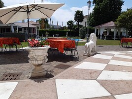 An outdoor dining area with tables covered by orange tablecloths, a large white umbrella, and a white lion statue.