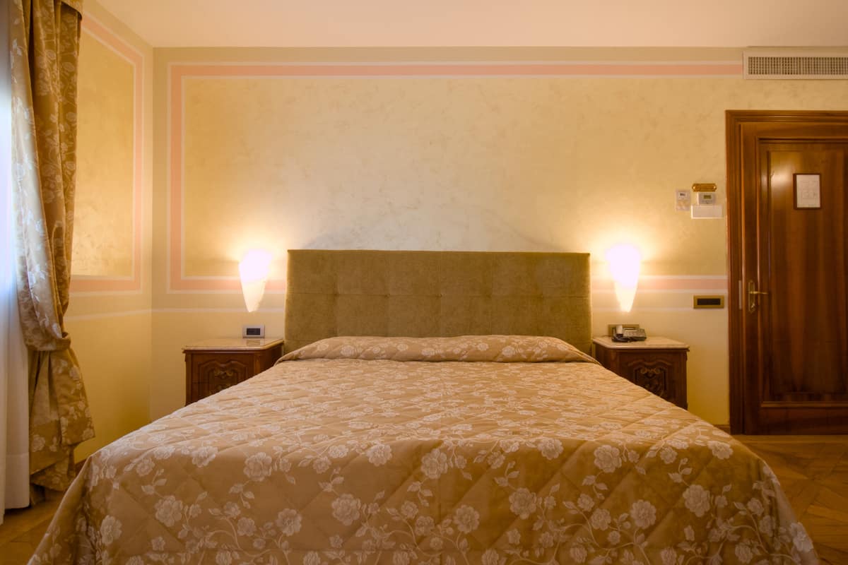 A hotel room with a large bed, two bedside tables, a wooden door, and a wall-mounted thermostat.