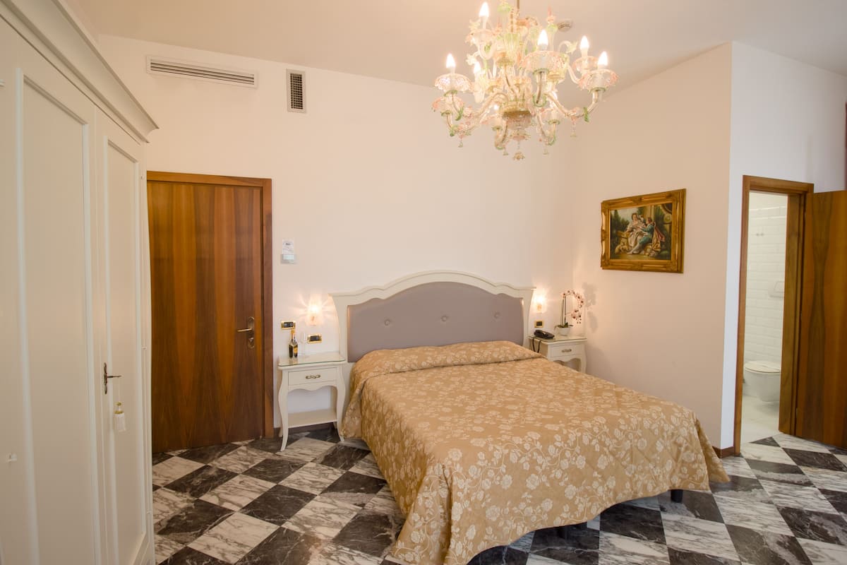 A bedroom with a checkered floor, a bed with a floral bedspread, a chandelier, a nightstand with a lamp, and a painting on the wall.