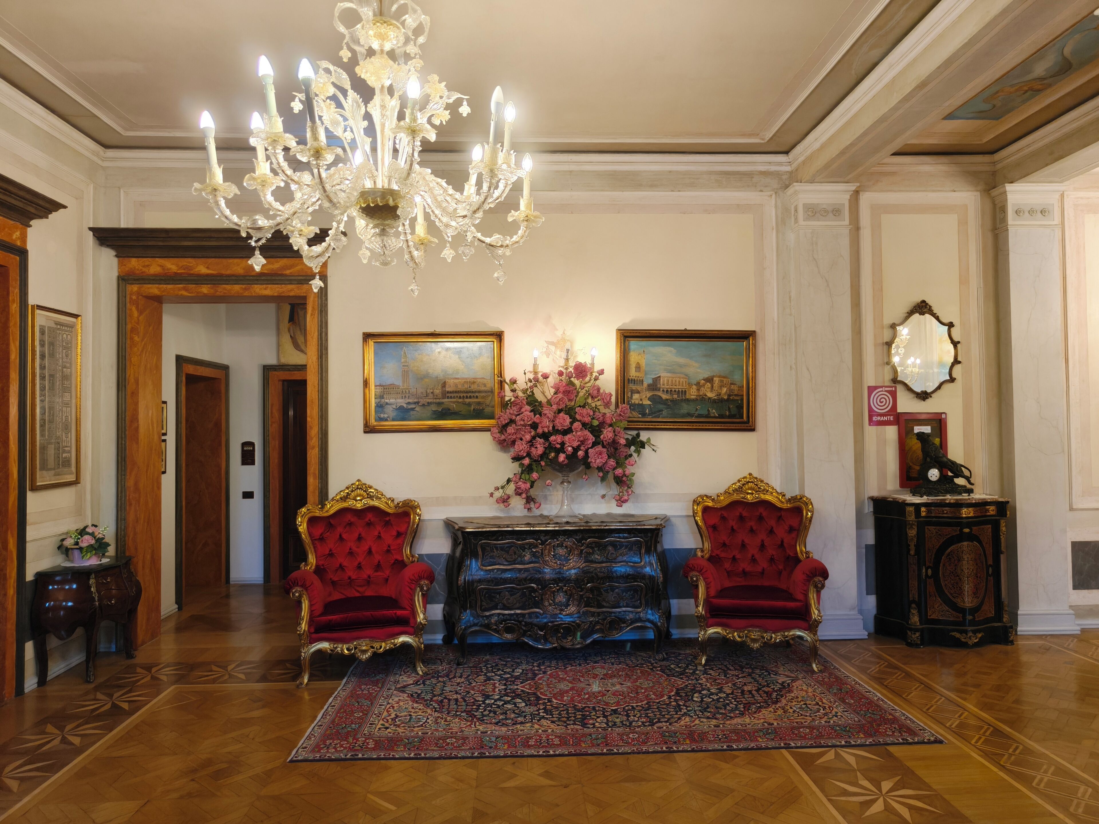 A grand lobby with a chandelier, red upholstered chairs, a console table, and a patterned rug.