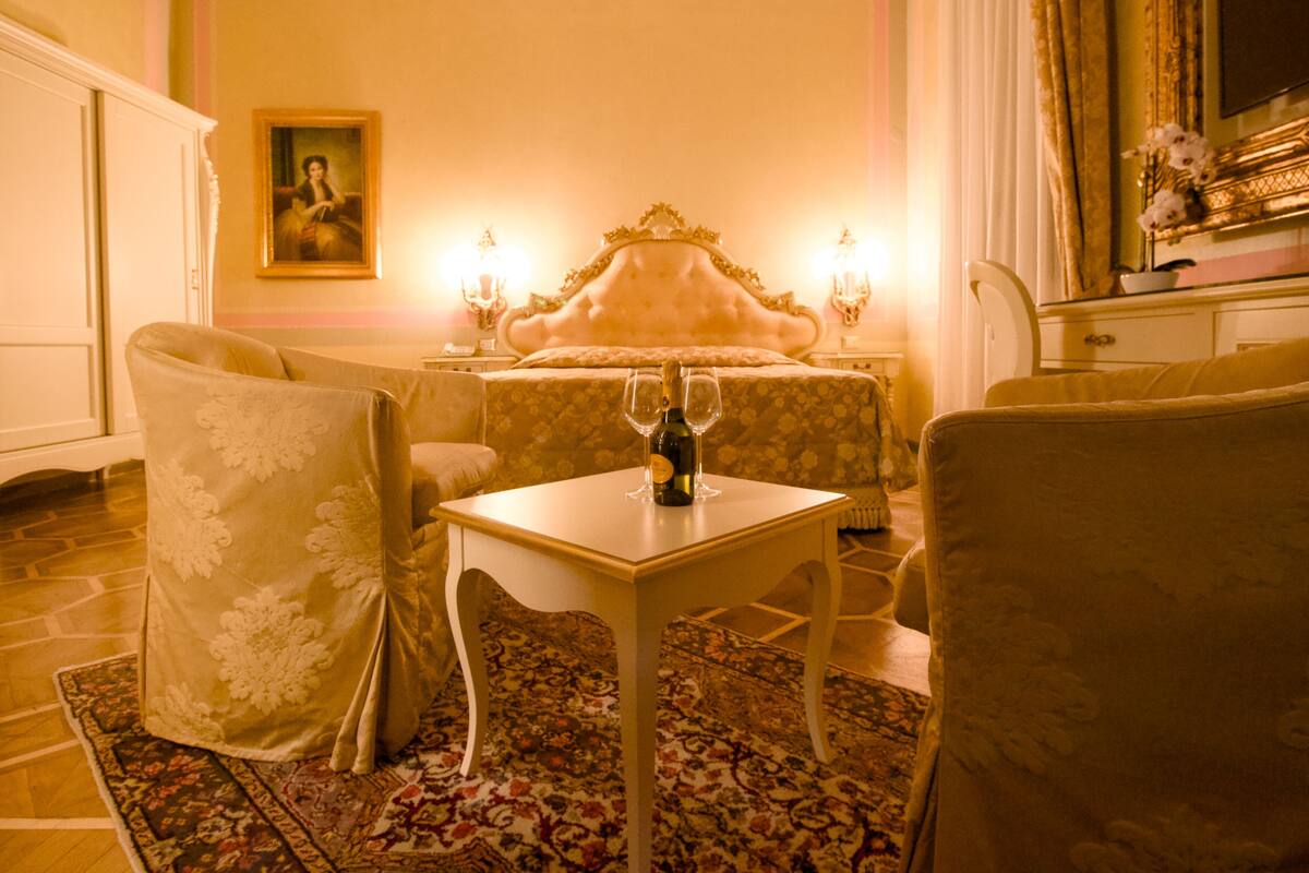 A room with a bed, two chairs, a table with a bottle and glasses, a lamp, and a painting on the wall.